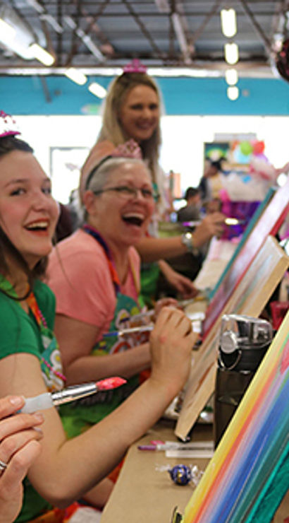 Smiling women at a lively indoor paint party in an art studio, wearing tiaras and aprons while painting colorful rainbow canvases on easels