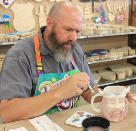 Bearded man in a colorful apron painting a Viking-themed ceramic mug at a DIY pottery-painting craft studio with shelves of wooden cutouts.