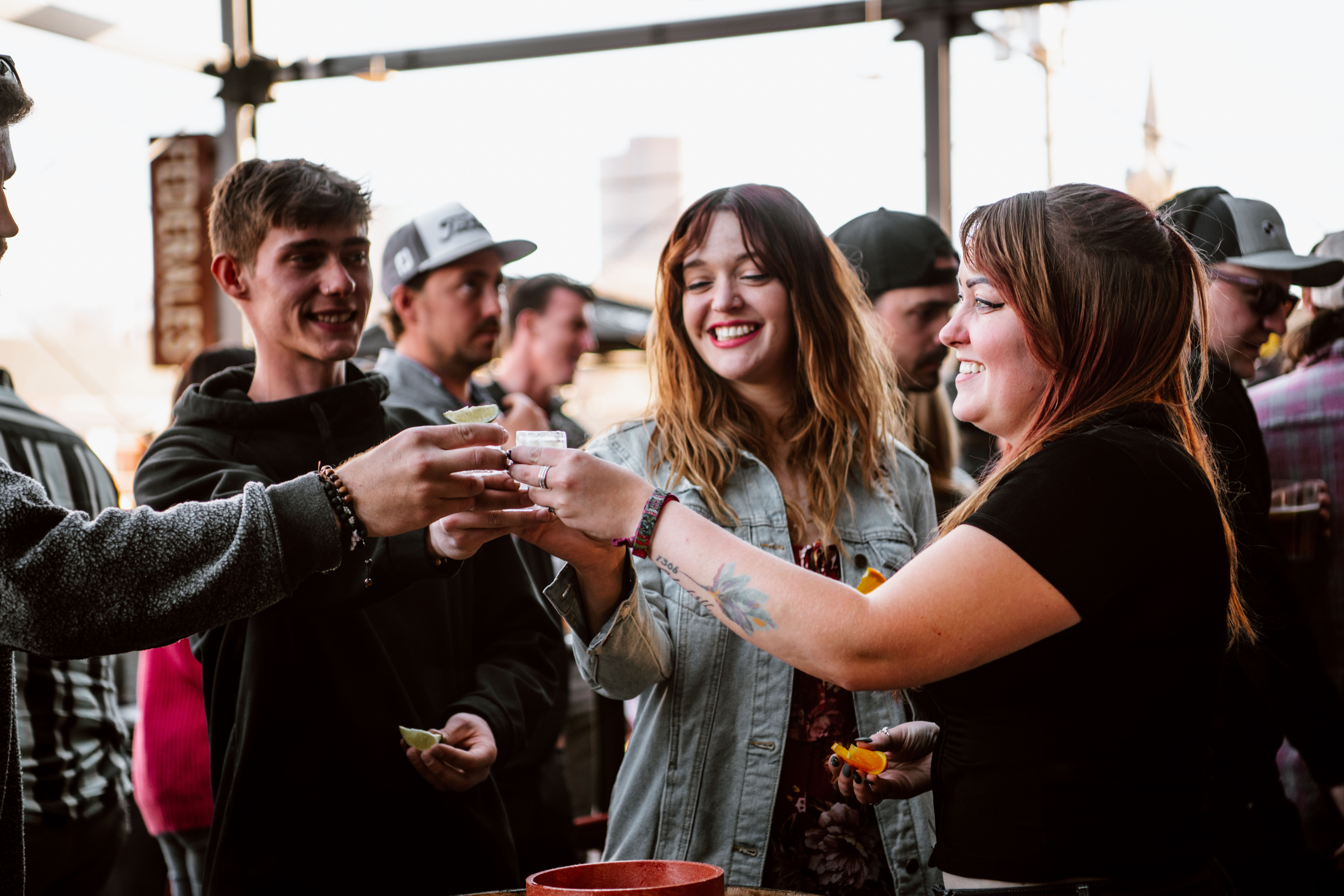 Smiling friends cheering as they clink shot glasses with lime and orange wedges on an outdoor bar patio, enjoying a lively nightlife gathering.