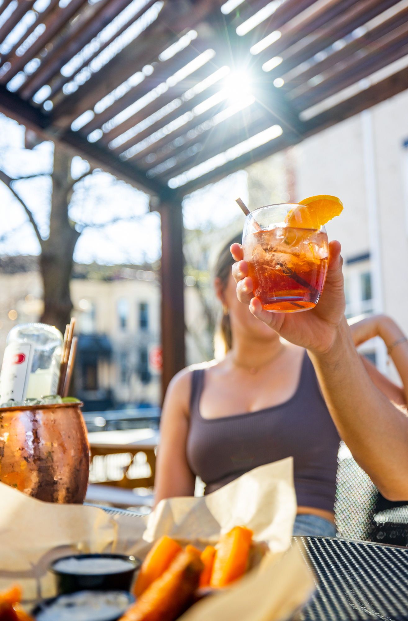 Hand holding an iced orange cocktail garnished with an orange slice on a sunny outdoor patio, with a copper drink bucket and fries with dipping sauces in a lively city brunch scene.