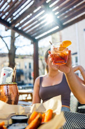 Hand holding an iced orange cocktail garnished with an orange slice on a sunny outdoor patio, with a copper drink bucket and fries with dipping sauces in a lively city brunch scene.