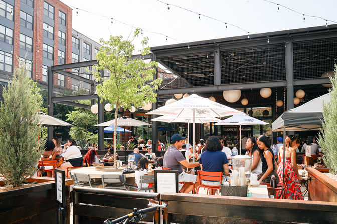 Bustling sunny urban outdoor patio with people dining under umbrellas and string lights in a modern industrial courtyard framed by trees and a brick building