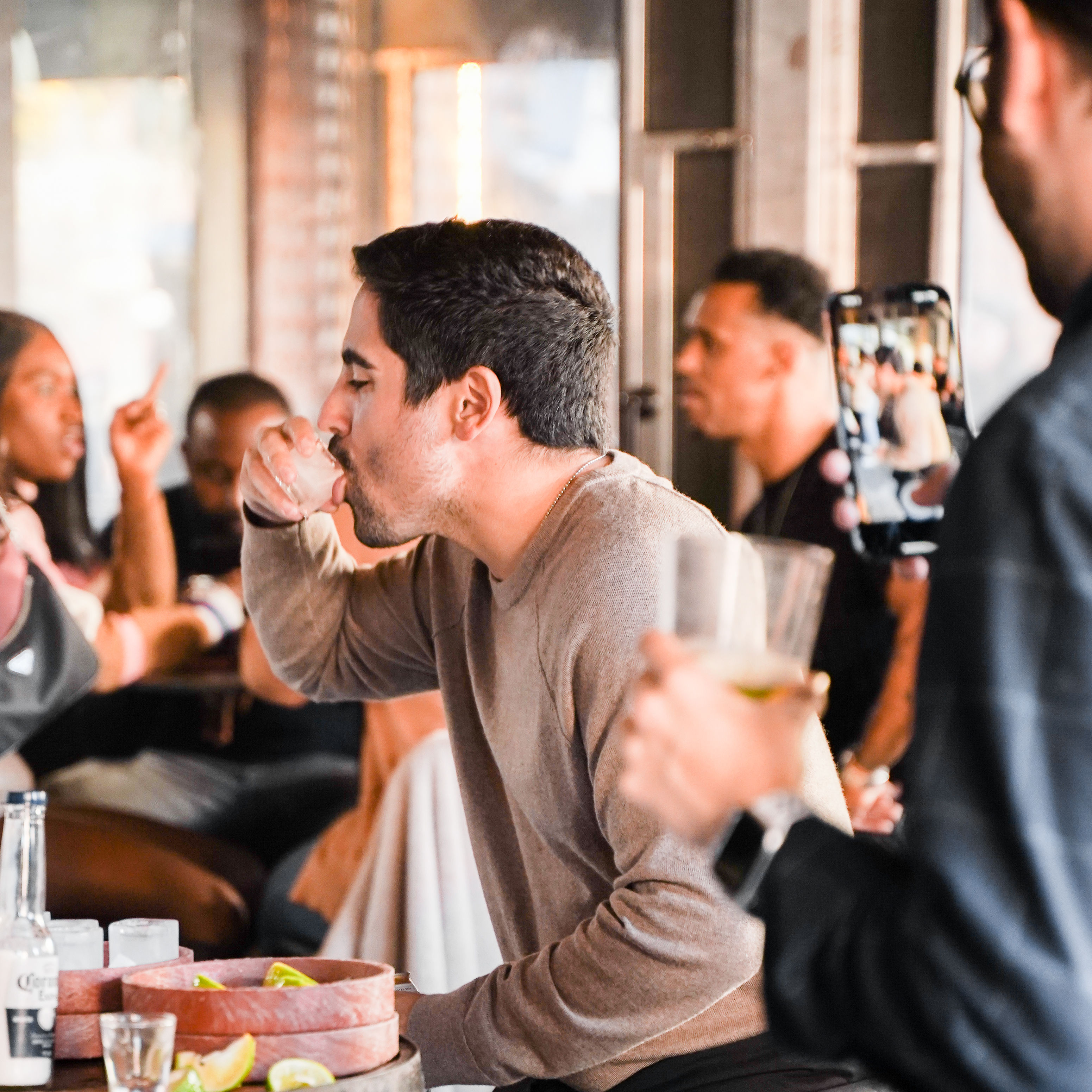 Man taking a shot at a lively urban bar with friends; someone films on a smartphone, lime wedges and beer bottles on the table, warm indoor lighting