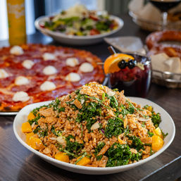 Hearty kale and farro salad with orange segments, toasted almonds and crunchy toppings on a restaurant table, with pepperoni pizza and a glass of sangria in the background.