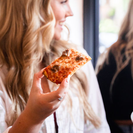 Close-up of a person holding a crispy rectangular cheese pizza slice in a sunlit cafe, smiling and chatting.