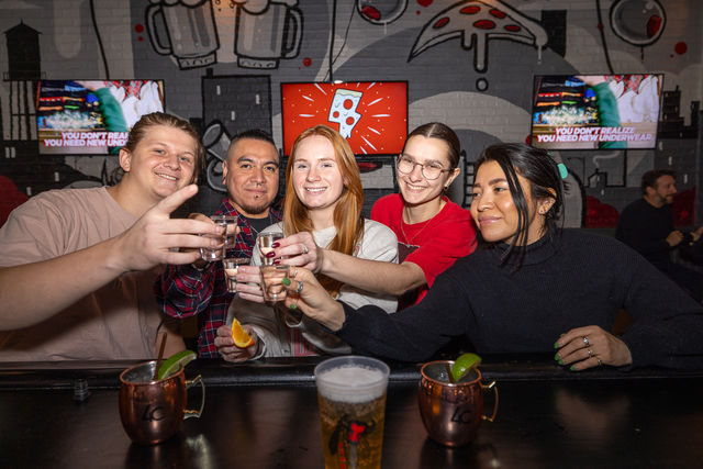Group of five friends at a lively bar raising shot glasses for a toast, with cocktails and beer on the counter and a colorful brick-wall mural featuring pizza and beer icons in the background.
