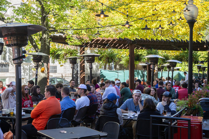 Bustling outdoor patio restaurant in autumn, groups dining under a wooden pergola with string lights, patio heaters and golden trees in an urban setting.