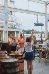 Friends celebrating on an urban rooftop patio bar at dusk — wooden barrels, high tables, casual summer outfits and cowboy boots with city buildings and lights in the background.