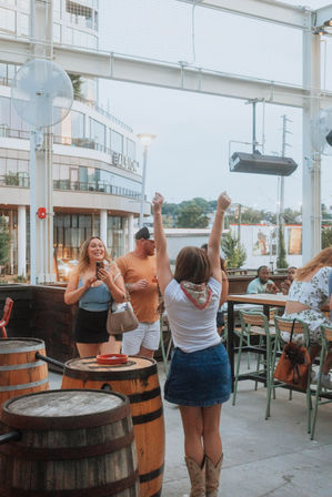 Friends celebrating on an urban rooftop patio bar at dusk — wooden barrels, high tables, casual summer outfits and cowboy boots with city buildings and lights in the background.