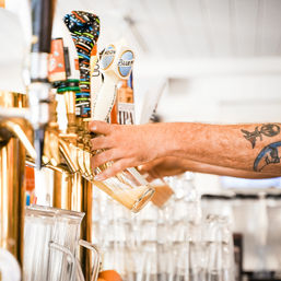 Tattooed arm pouring frothy draft beer from brass beer taps into a glass at a bright neighborhood bar with stacked glassware
