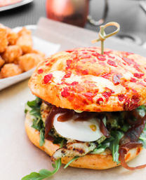 Close-up gourmet grilled chicken sandwich on a sun-dried tomato bun with arugula, fresh mozzarella, caramelized onions and a toothpick skewer, with crispy tater tots blurred in the background.