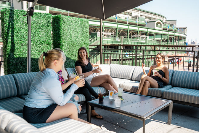 Four friends enjoying drinks on a sunny rooftop patio lounge with striped cushions and umbrella, decorative green hedge panels and a large green stadium/ballpark visible in the background.