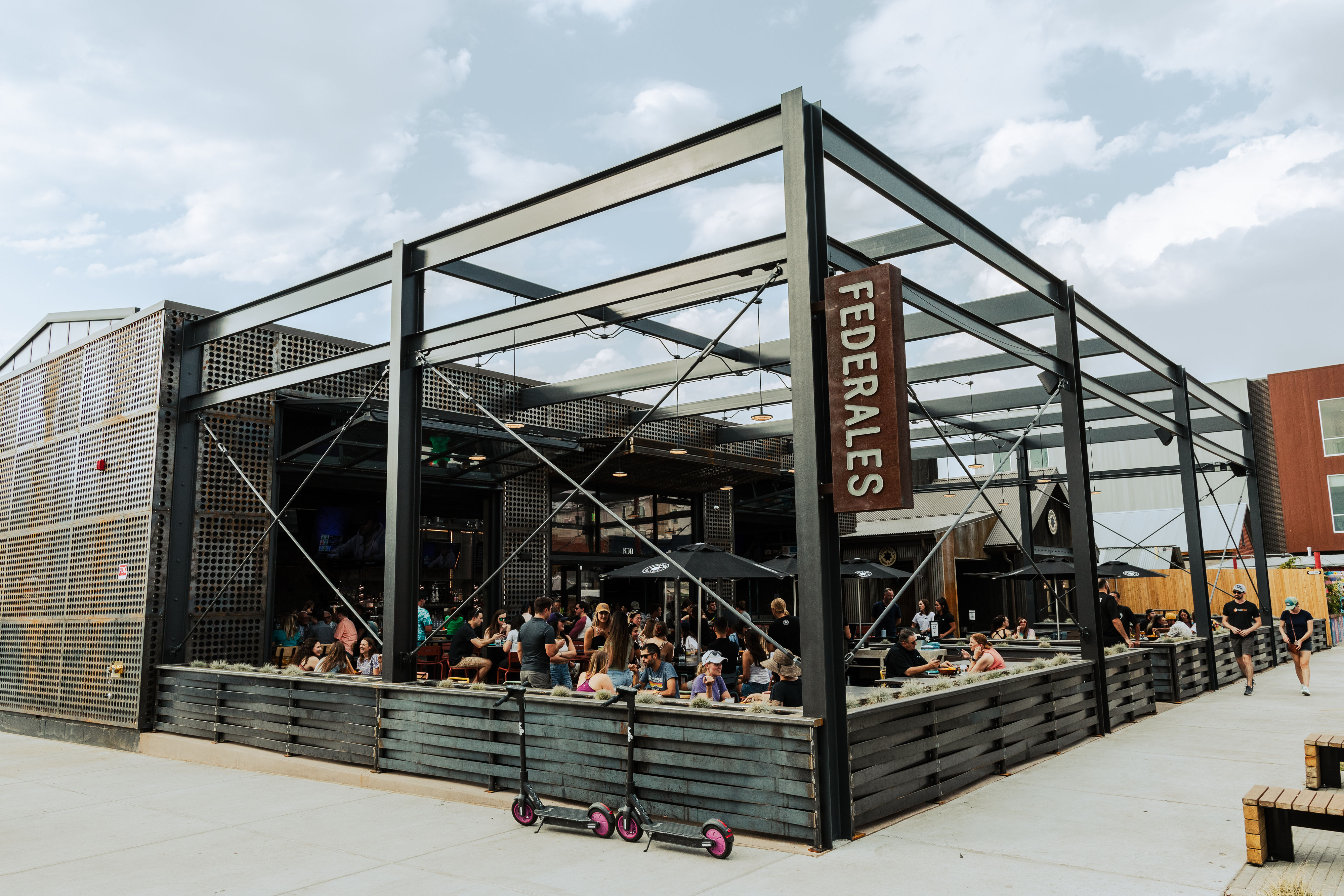 Busy outdoor urban patio with black steel-frame pergola and fenced seating area, people dining under umbrellas on a sunny day and parked electric scooters along the sidewalk.