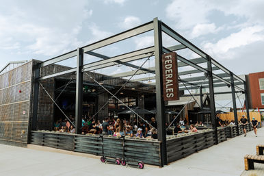 Busy outdoor urban patio with black steel-frame pergola and fenced seating area, people dining under umbrellas on a sunny day and parked electric scooters along the sidewalk.