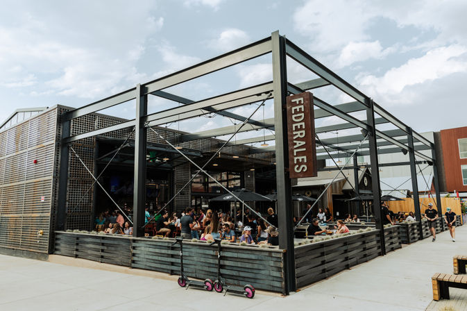 Busy outdoor urban patio with black steel-frame pergola and fenced seating area, people dining under umbrellas on a sunny day and parked electric scooters along the sidewalk.