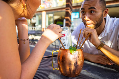 Sunlit outdoor patio scene of two people sharing a large copper cocktail mug with lime and an inverted can, sipping through multiple straws