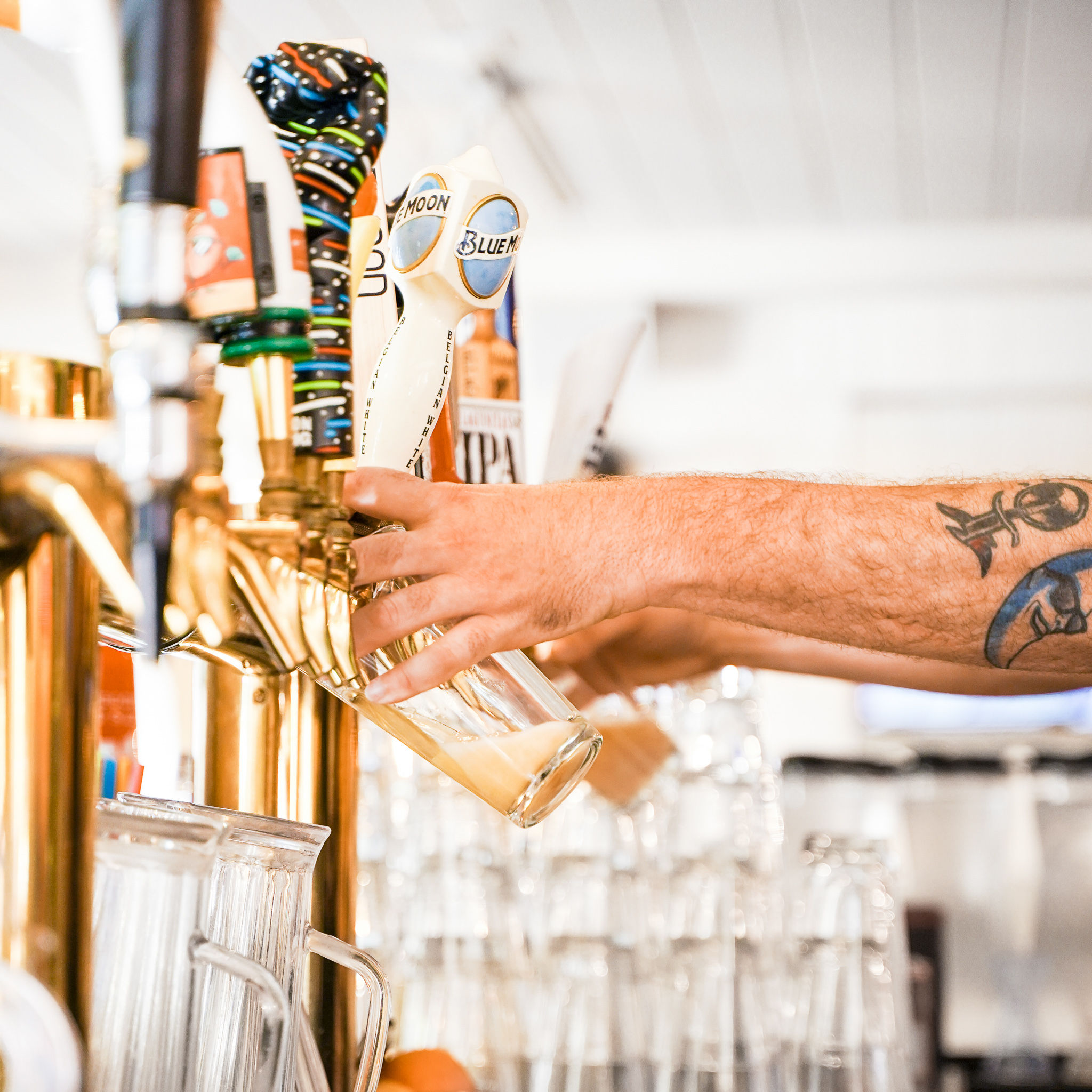 Tattooed bartender pouring craft draft beer from brass tap handles into a tilted pint glass at a bright bar with stacked glassware