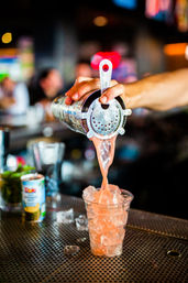 Close-up of a bartender pouring a pink cocktail from a metal shaker through a strainer into an ice-filled plastic cup on a busy bar counter, vibrant craft cocktail scene