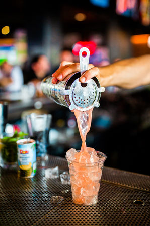 Close-up of a bartender pouring a pink cocktail from a metal shaker through a strainer into an ice-filled plastic cup on a busy bar counter, vibrant craft cocktail scene