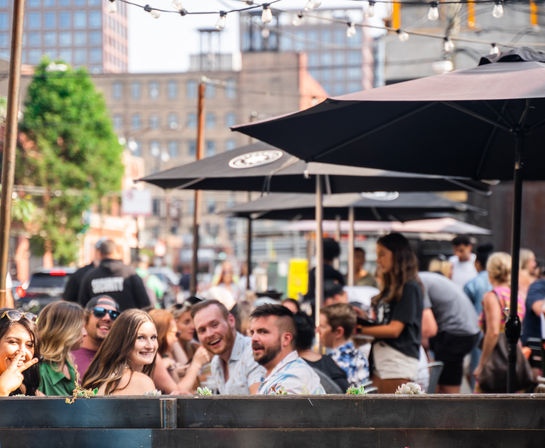 Smiling group of friends enjoying al fresco dining on a busy downtown urban patio with black umbrellas, string lights, and city buildings in the background.