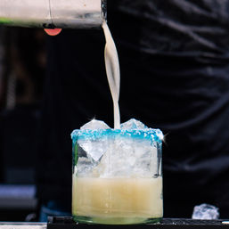 Close-up of a creamy cocktail being poured into an ice-filled glass with a bright blue salt rim, bartender in background