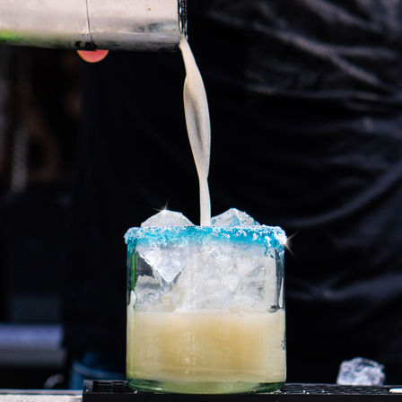 Close-up of a creamy cocktail being poured into an ice-filled glass with a bright blue salt rim, bartender in background