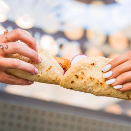 Close-up of two hands holding soft street-style tacos with radish slices and crispy filling against bokeh market lights