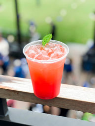 Bright pink iced drink topped with a mint leaf in a clear plastic cup resting on a wooden railing, blurred outdoor sports field and crowd in the background
