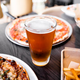 Pint of amber craft beer on a dark wood table surrounded by thin-crust margherita pizzas and a tray of fries at a casual pizzeria