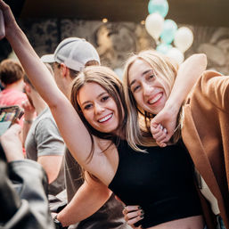 Two friends smiling and hugging at a lively indoor party with teal and white balloons — fun city nightlife celebration