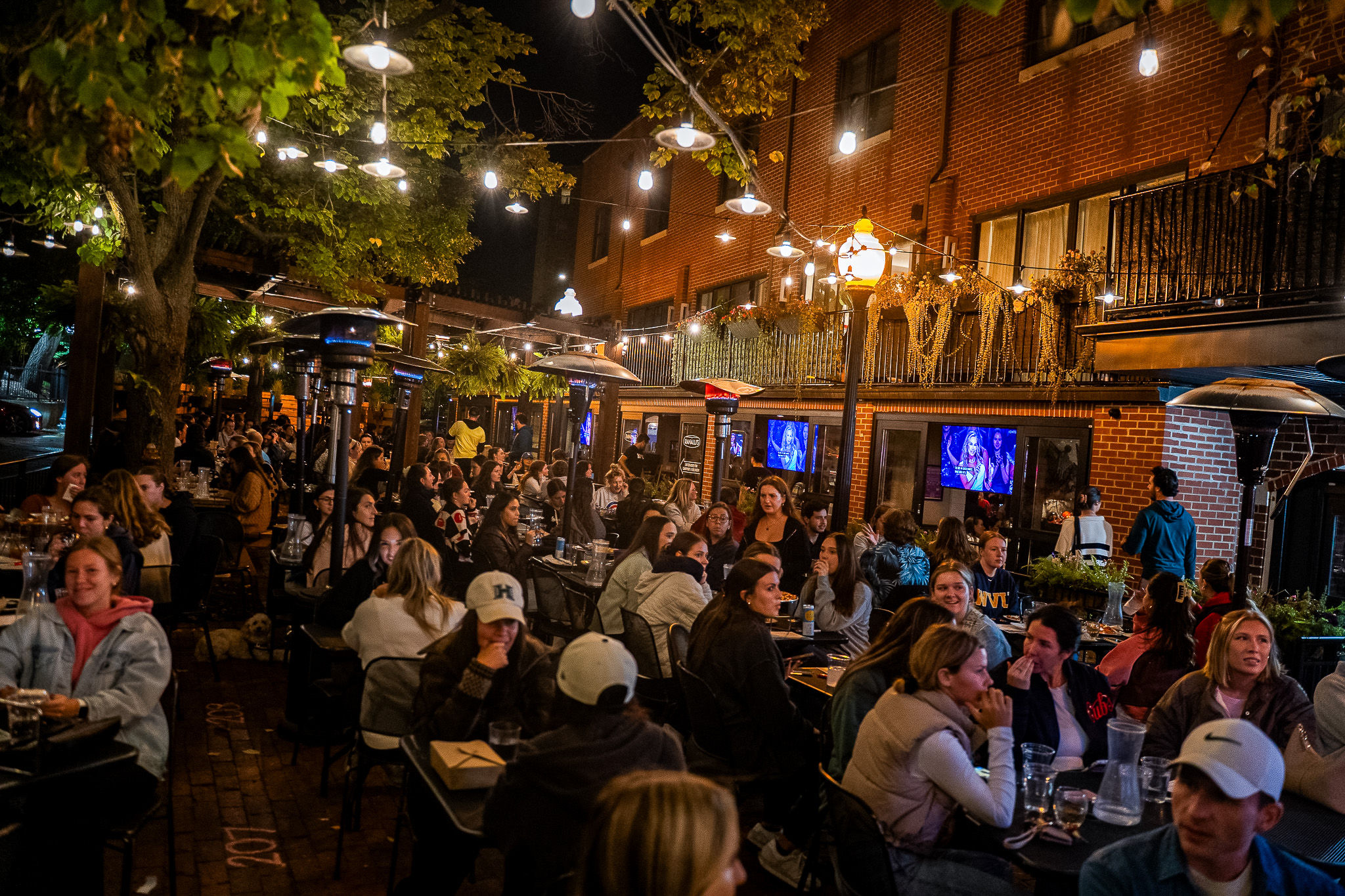Lively downtown outdoor restaurant patio at night — crowded al fresco dining under string lights, patio heaters, and brick storefronts.
