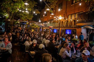 Lively downtown outdoor restaurant patio at night — crowded al fresco dining under string lights, patio heaters, and brick storefronts.