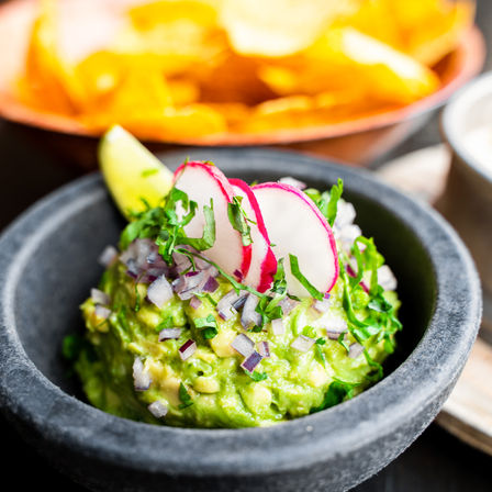 Bright Mexican-style guacamole topped with diced red onion, chopped cilantro and radish slices with a lime wedge in a stone bowl, blurred tortilla chips in the background