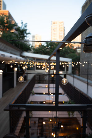Cozy downtown urban rooftop patio at dusk, warm bistro string lights draped across a metal pergola over a multi-level outdoor dining area with blurred city skyline in the background.