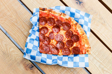 Pepperoni pizza slice on blue-and-white checkered paper atop a wooden picnic table, golden crust and melted cheese close-up.