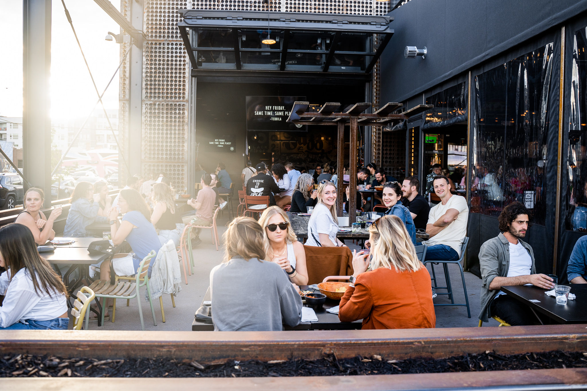 Sunlit downtown outdoor patio with groups of people dining al fresco at city restaurant tables, lively casual atmosphere and friends chatting.