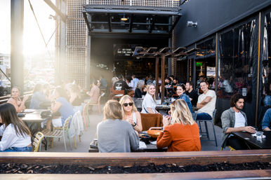 Sunlit downtown outdoor patio with groups of people dining al fresco at city restaurant tables, lively casual atmosphere and friends chatting.