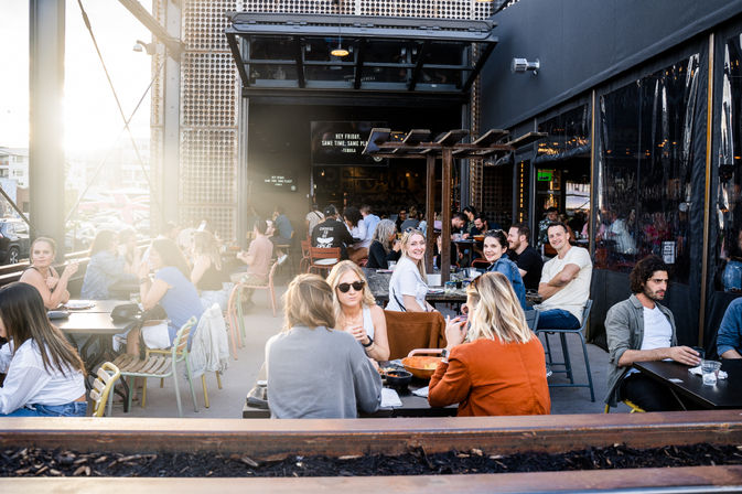 Sunlit downtown outdoor patio with groups of people dining al fresco at city restaurant tables, lively casual atmosphere and friends chatting.