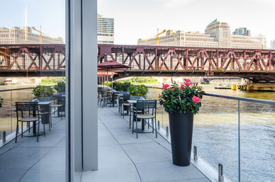 Sunny riverside café terrace with empty tables and tall planters of pink flowers, glass railing overlooking the Chicago River and a rust-red steel bridge with downtown buildings beyond.