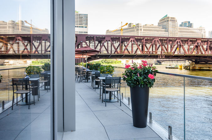 Sunny riverside café terrace with empty tables and tall planters of pink flowers, glass railing overlooking the Chicago River and a rust-red steel bridge with downtown buildings beyond.