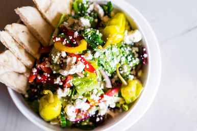 Overhead close-up of a vibrant Mediterranean salad bowl with mixed greens, crumbled feta, sliced red peppers, cucumbers, red onion, zesty pepperoncini and pita wedges