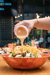 Hand pouring creamy chipotle sauce over loaded nachos topped with crispy tortilla strips, cilantro and cheese in a wooden bowl on a restaurant patio