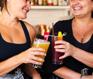 Two friends laughing and clinking drinks at a bar — one holding a hazy craft beer and the other a blackberry‑orange cocktail with garnish.