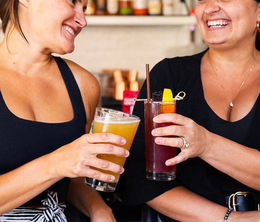 Two friends laughing and clinking drinks at a bar — one holding a hazy craft beer and the other a blackberry‑orange cocktail with garnish.