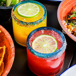 Vibrant yellow and red margaritas in blue-rimmed glasses with lime wheel garnish and salt-and-chili rims on a restaurant table with chips and bowls