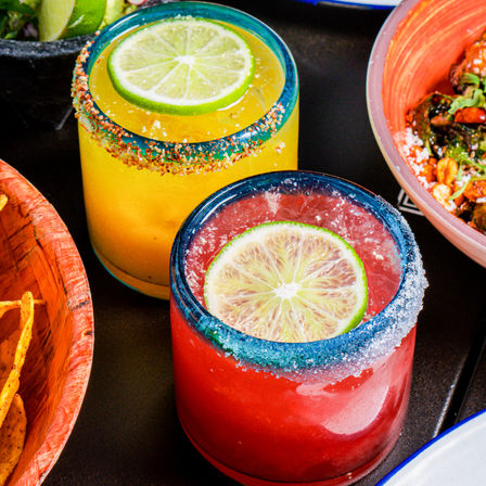 Vibrant yellow and red margaritas in blue-rimmed glasses with lime wheel garnish and salt-and-chili rims on a restaurant table with chips and bowls