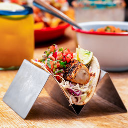 Grilled fish taco with charred fillet, fresh pico de gallo, shredded cabbage and lime in a soft tortilla, held in a metal taco stand on a wooden table