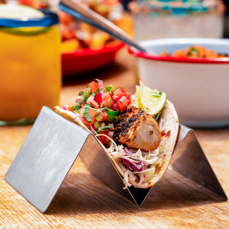 Grilled fish taco with charred fillet, fresh pico de gallo, shredded cabbage and lime in a soft tortilla, held in a metal taco stand on a wooden table
