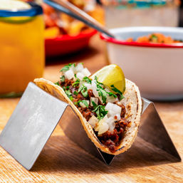 Authentic Mexican street-style corn taco with seasoned meat, chopped onions, cilantro and a lime wedge in a metal taco holder on a wooden table, blurred salsa and drink in the background.