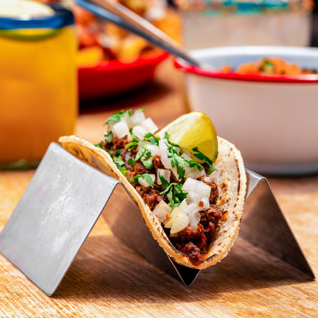 Authentic Mexican street-style corn taco with seasoned meat, chopped onions, cilantro and a lime wedge in a metal taco holder on a wooden table, blurred salsa and drink in the background.